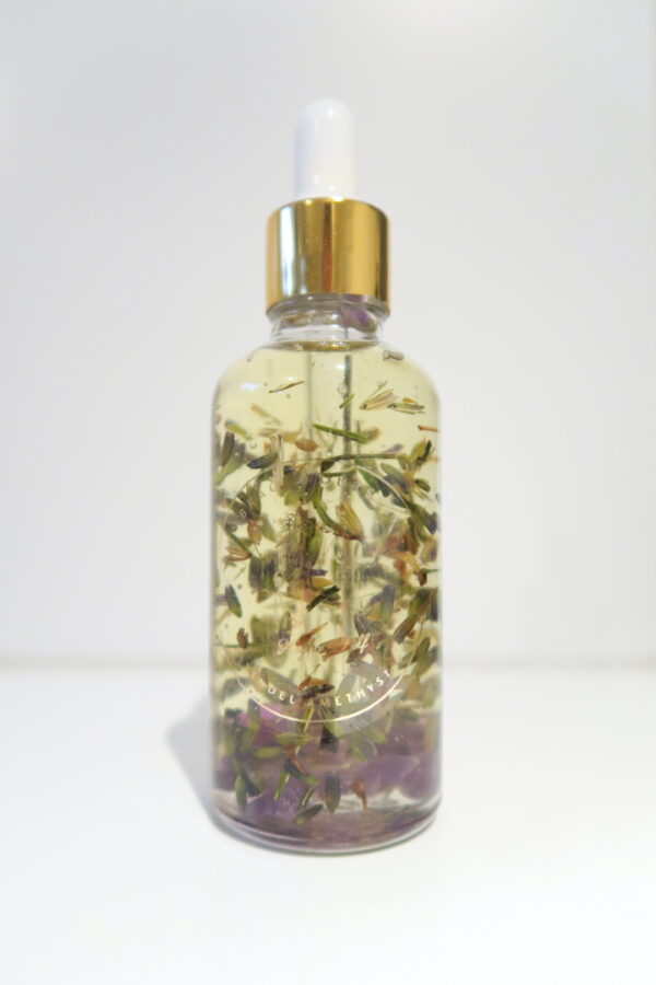 Een flesje verzorgende amethist gezichts- en lichaamsolie, omringd door lavendelbloemen en amethiststenen.