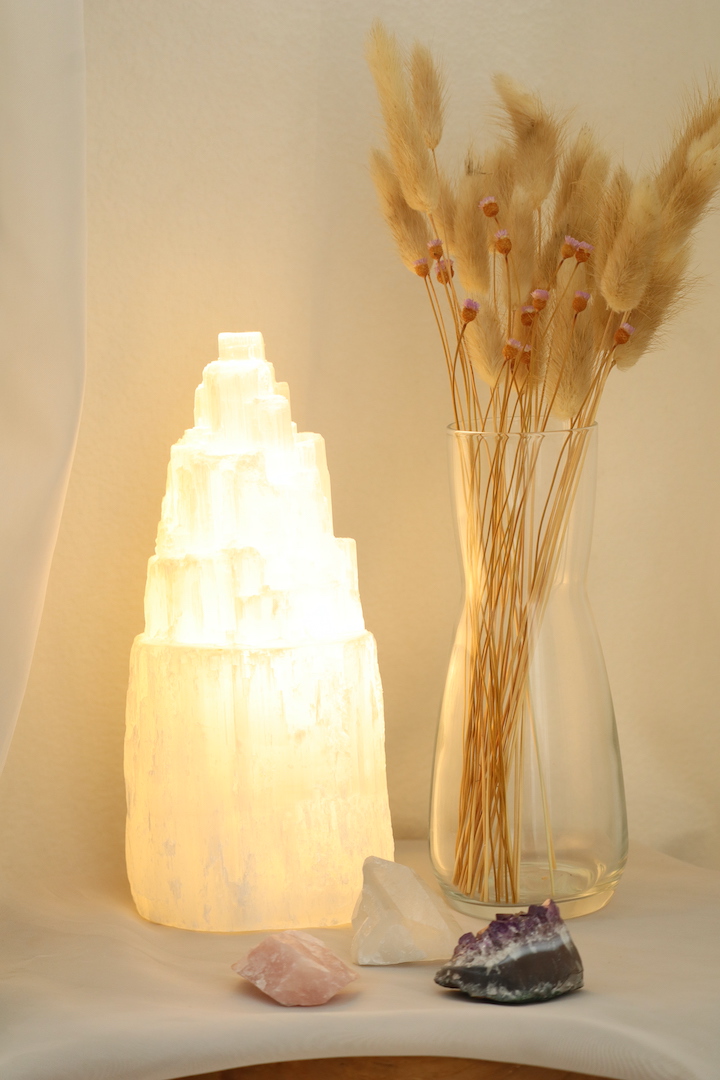 Seleniet lamp van circa 21 cm hoog met warm wit licht, naast een glazen vaas met droogbloemen en drie edelstenen op een wit kleed.