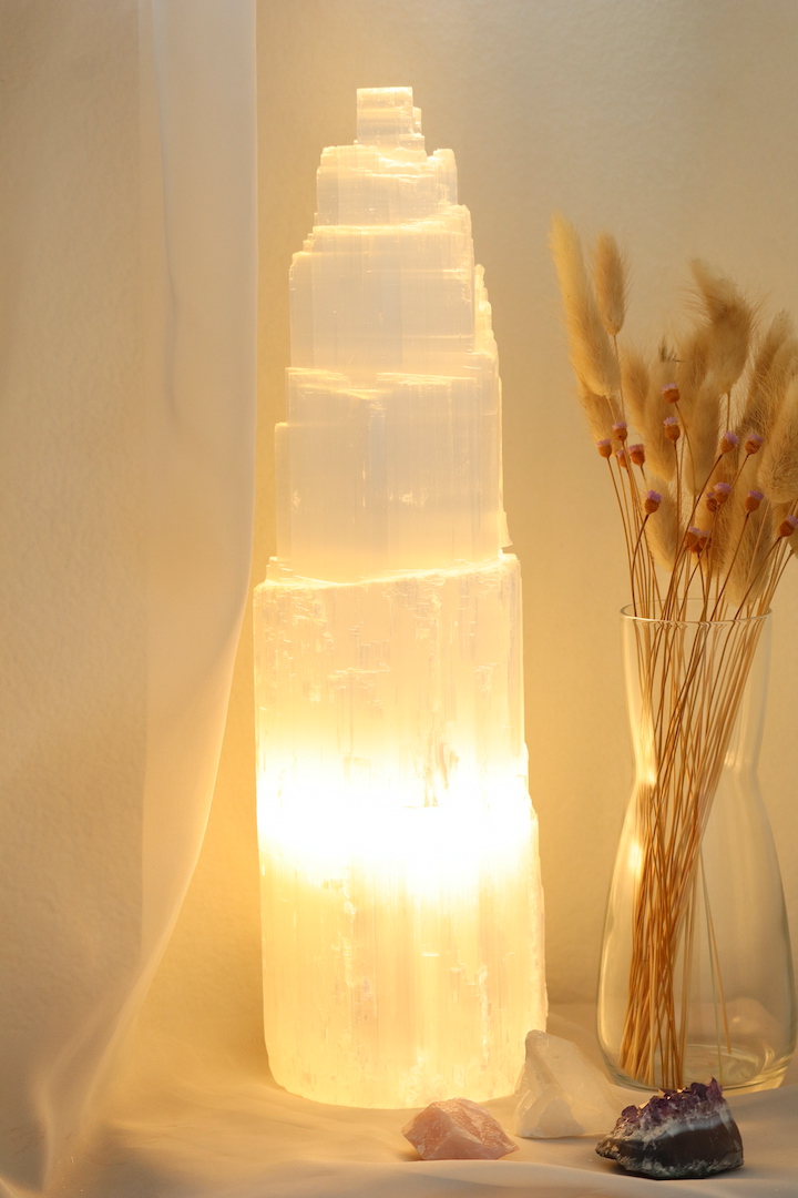 Grote Seleniet lamp van circa 35 cm hoog met warme witte gloed, naast glazen vaas met droogbloemen en edelstenen op houten tafel.