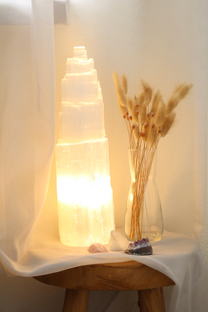 Grote Seleniet lamp van circa 35 cm hoog met warme witte gloed, naast glazen vaas met droogbloemen en edelstenen op houten tafel.