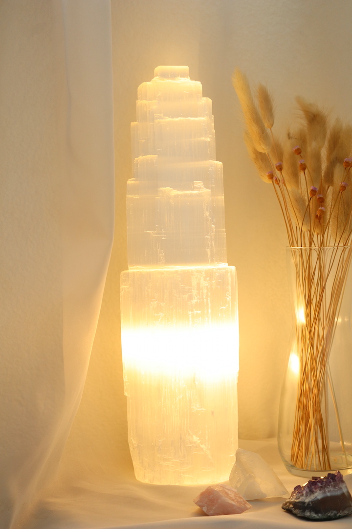 Grote Seleniet lamp van circa 35 cm hoog met warme witte gloed, naast glazen vaas met droogbloemen en edelstenen op houten tafel.