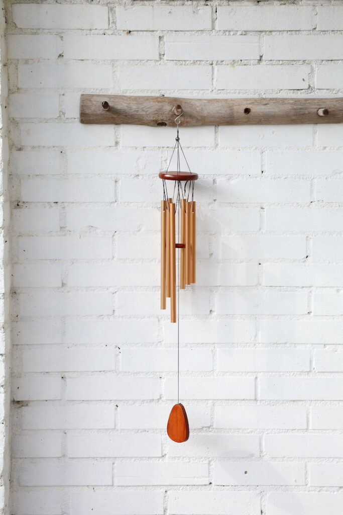 Rosékleurige windgong van 68 cm lang en 8,5 cm breed met houten details, hangend aan een witte muur in zacht zonlicht.