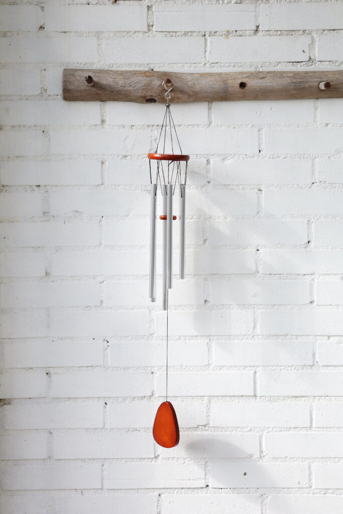 Zilverkleurige windgong van 68 cm lang en 8,5 cm breed met houten details, hangend aan een witte muur in het zonlicht.