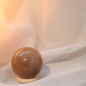 AA-kwaliteit Perzik Maansteen met Zonnesteen bol van 4,5 cm met sterke gouden sparkle, gefotografeerd in warm zacht licht op een lichte houten standaard.