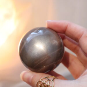Zwarte Perzik Maansteen met Zonnesteen bol van 5,2 cm met subtiele gouden shimmer, geplaatst op een houten standaard in warm diffuus licht.