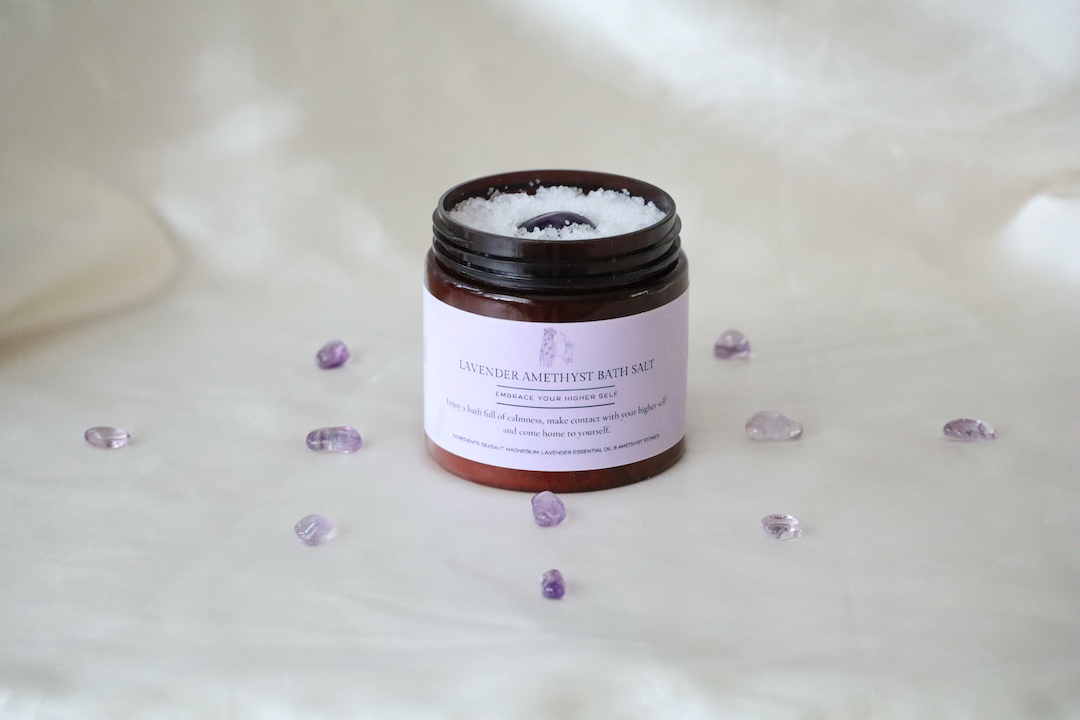 “Lavendel Amethist badzout in amberkleurige pot met amethist steentjes en lavendelaccenten, naast een maatlepel gevuld met badzout.”