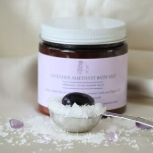 “Lavendel Amethist badzout in amberkleurige pot met amethist steentjes en lavendelaccenten, naast een maatlepel gevuld met badzout.”
