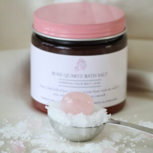“Amberkleurige pot Rozenkwarts Roos Badzout met Himalayazout, rozenkwarts trommelsteen en kleine rozenkwarts steentjes, naast een maatlepel met badzout.”