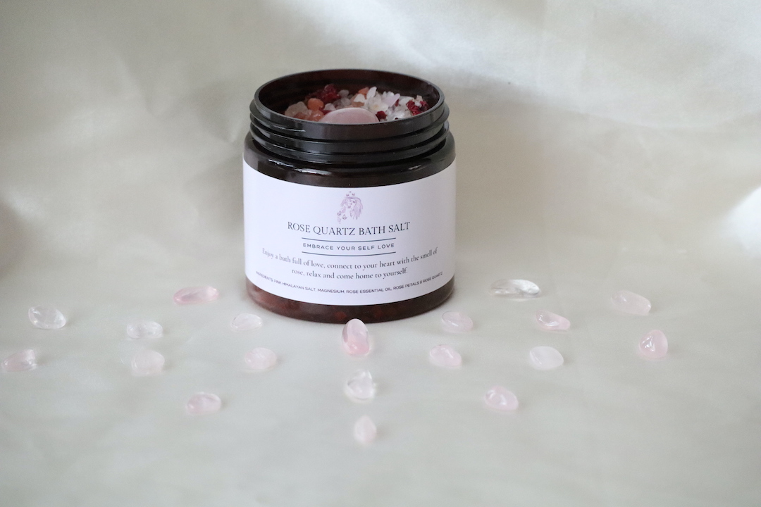 “Amberkleurige pot Rozenkwarts Roos Badzout met Himalayazout, rozenkwarts trommelsteen en kleine rozenkwarts steentjes, naast een maatlepel met badzout.”