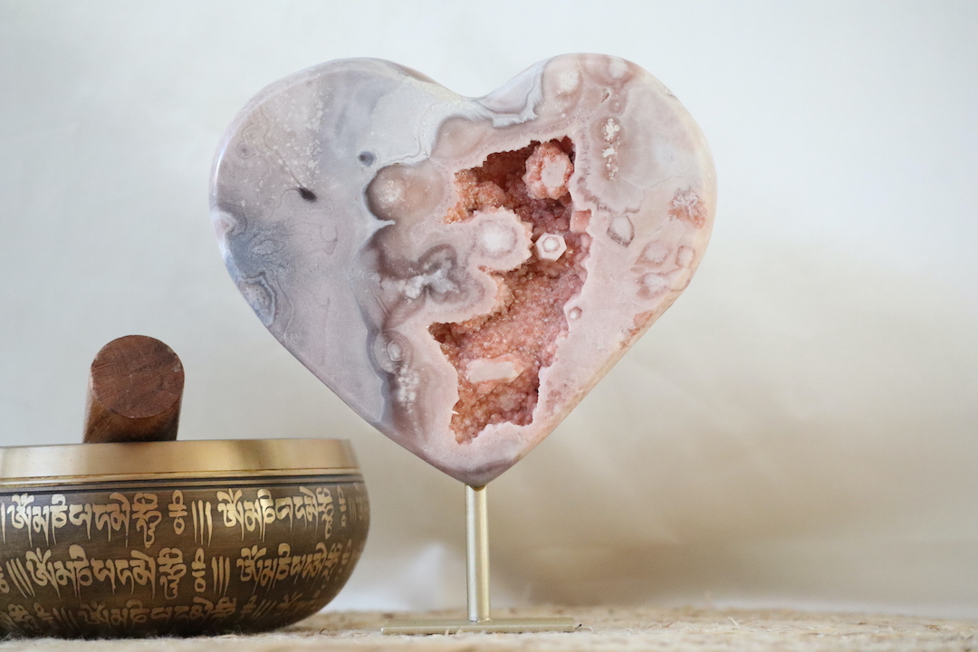 Druzy chalcedoon hart op metalen standaard met zachte beige en crème tinten en een open kristalholte, gefotografeerd in natuurlijk daglicht.