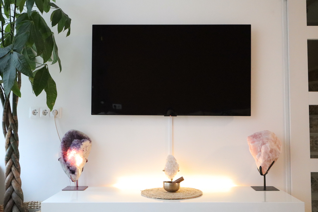 Gouden driehoek met verlichte amethist en rozenkwarts op standaard in een minimalistisch interieur, opgesteld op een witte kast onder een wandgemonteerde televisie.