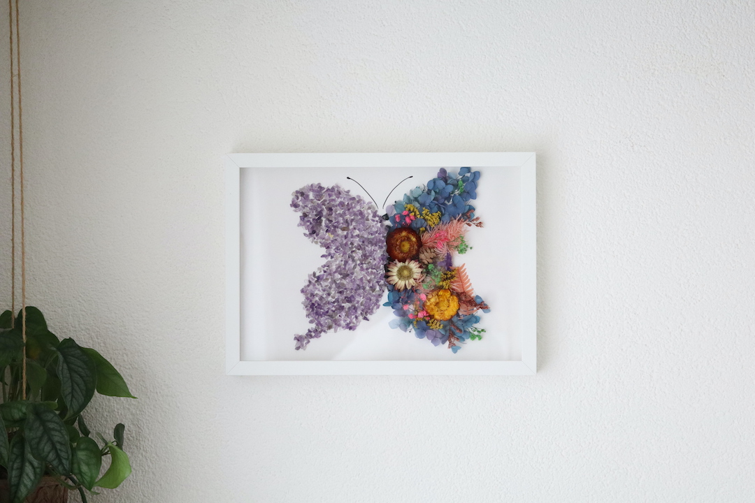 Handgemaakte vlinder wanddecoratie met amethist kristallen en gedroogde bloemen in witte lijst.