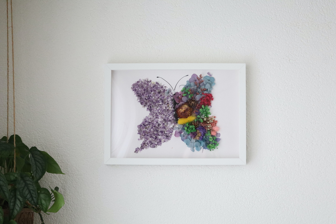 Handgemaakte vlinder wanddecoratie met amethist kristallen en gedroogde bloemen in witte lijst.