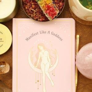 Roze Manifest Like a Goddess manifestatie journal met gouden godinnenillustratie en rozenkwarts pen, omringd door edelstenen en self-care accessoires.