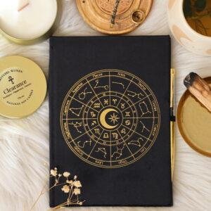 Zwart fluwelen sterrenbeeld notitieboekje met gouden astrologisch wiel en obsidiaan pen, omringd door kaars, wierook en kruidenthee.
