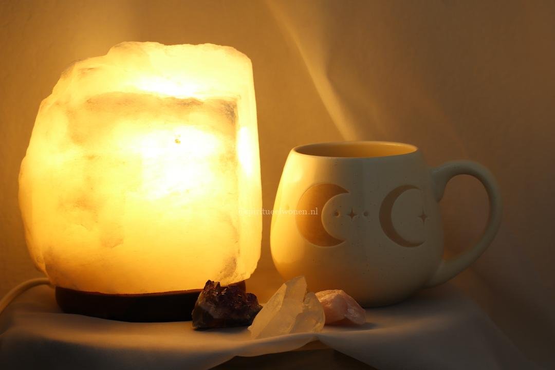 Witte zoutlamp met warme verlichting naast keramieken maanfasen mok, amethist, bergkristal en rozenkwarts.