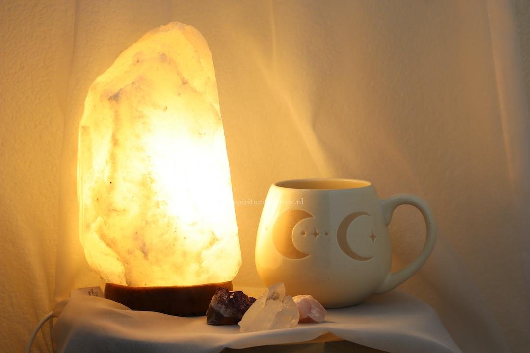 Witte zoutlamp met warme verlichting naast keramieken maanfasen mok, amethist, bergkristal en rozenkwarts.