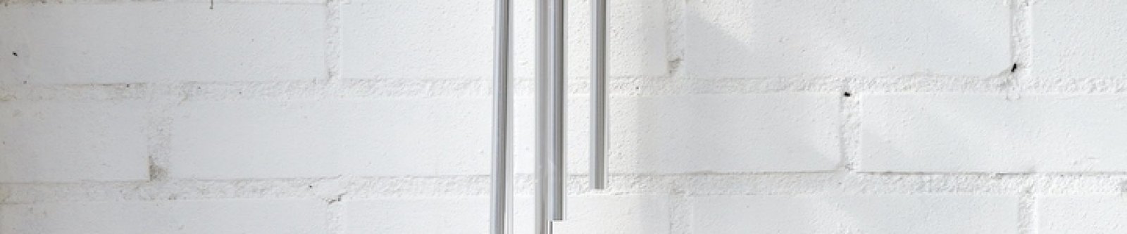 Zilverkleurige windgong van 68 cm lang en 8,5 cm breed met houten details, hangend aan een witte muur in het zonlicht.